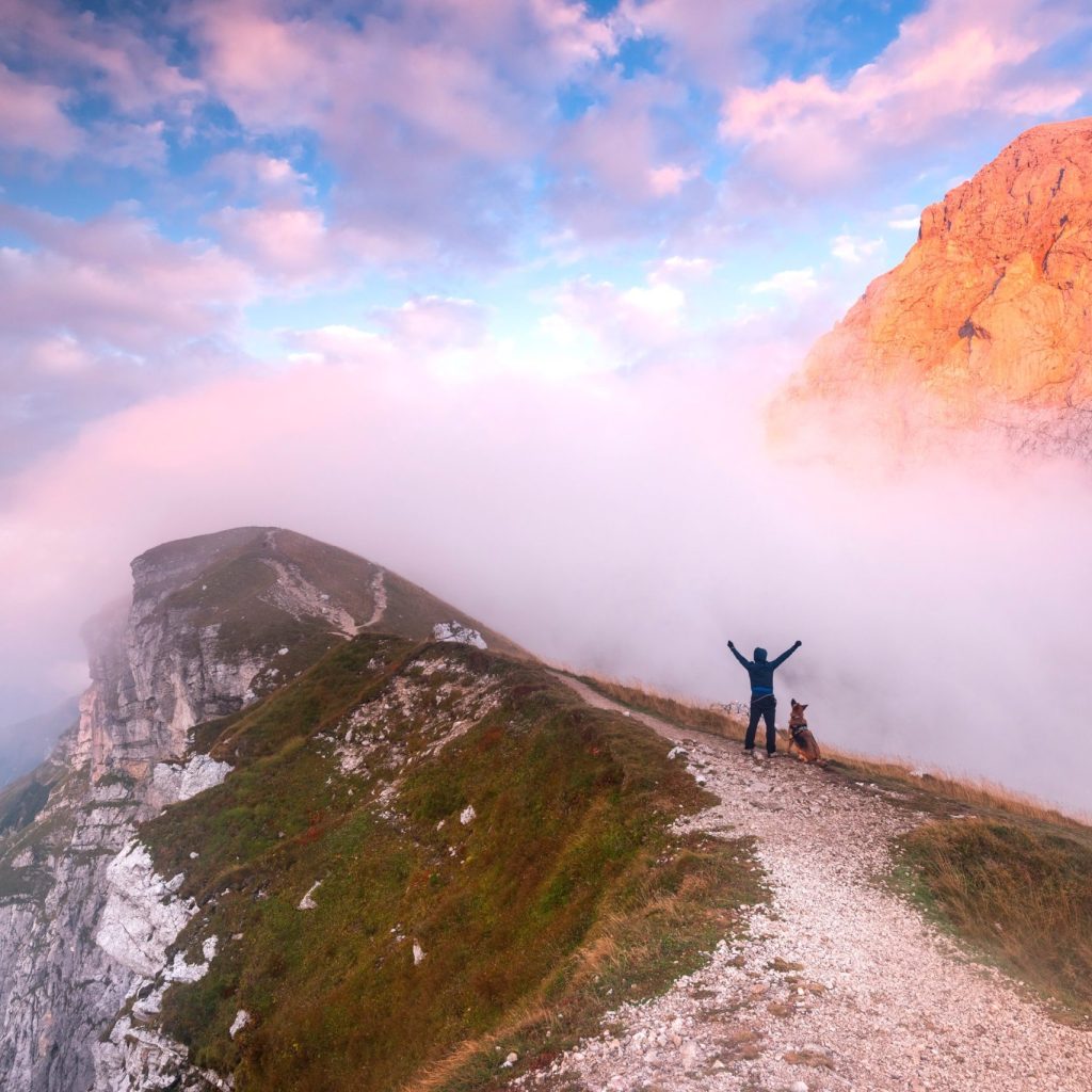 Adventure Man with Dog Standing at Mountain at Dramatic Sunset.